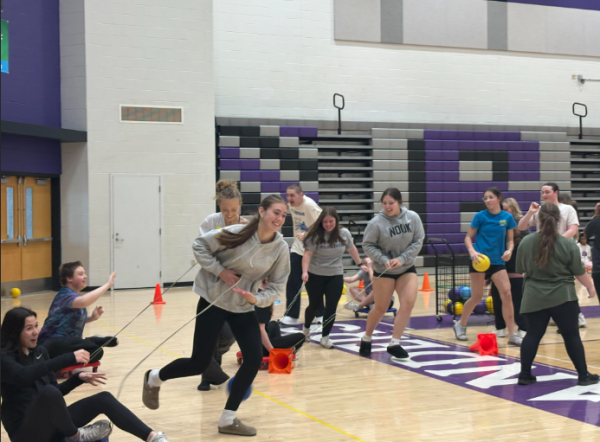 The photo shows about a dozen high school students playing in a gym. Some students sit on scooters with jump ropes looped around them. Other students hold onto the jump ropes to pull them around the gym for a racing game.