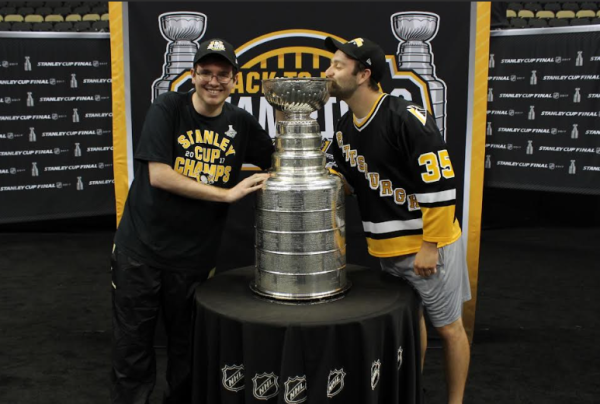 The photo shows Tommy Mowers and Anthony Barbano in Pittsburgh Penguins merchandise. They stand on either side of the Stanley Cup. Mowers is smiling at the camera and Barbano is kissing the Cup.