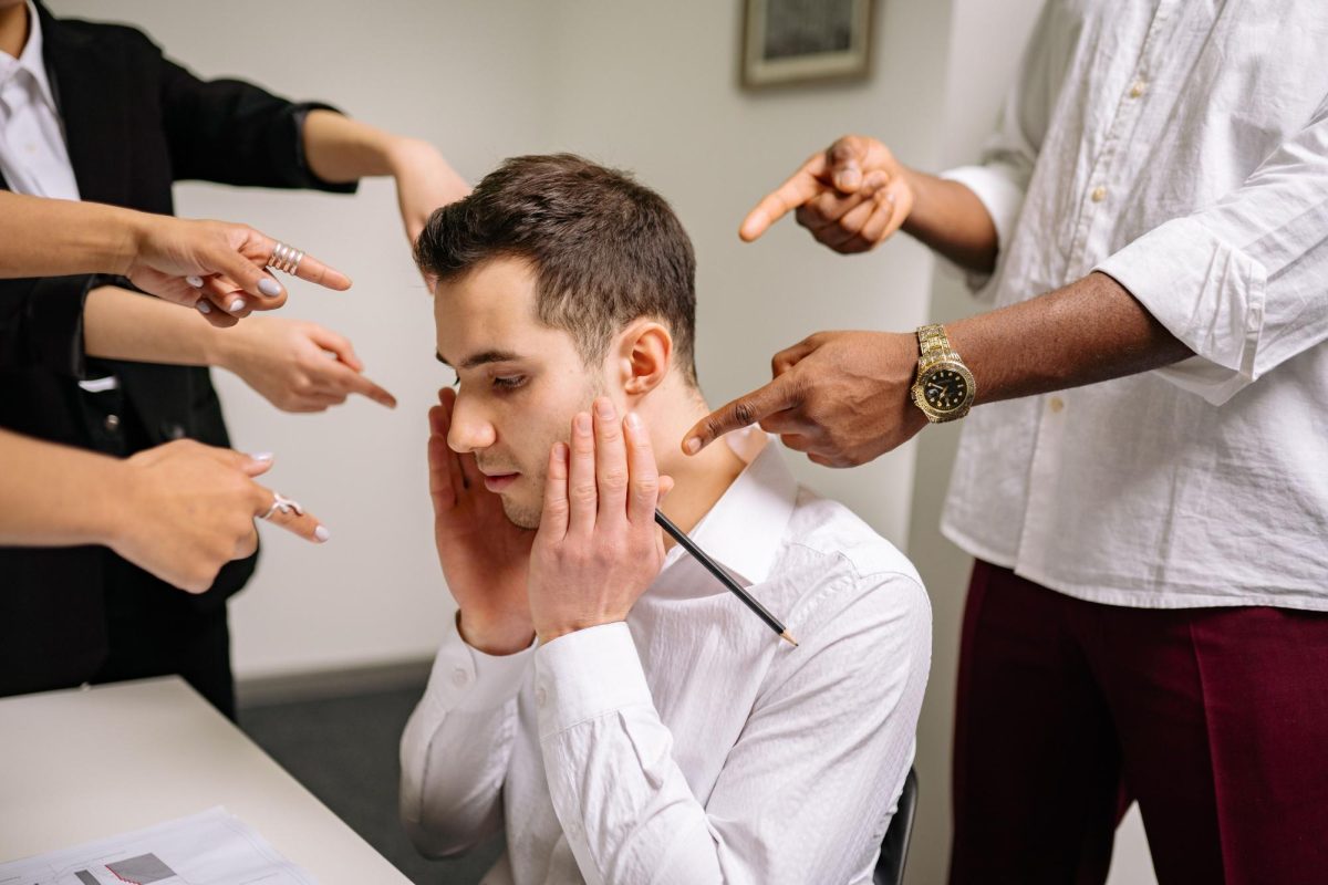 A stressed man at the office surrounded by pointing fingers, representing workplace bullying and stress. Image via Pexels